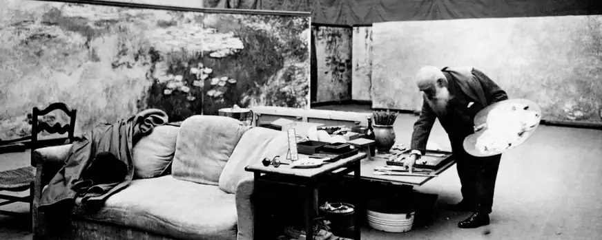 Black-and-white photograph of a painter holding a palette in a studio filled with canvases